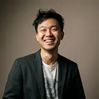 a12493ee-78f5-49d4-bd8c-000de4c777a3.Portrait of a man smiling in gray sweater against dark background.